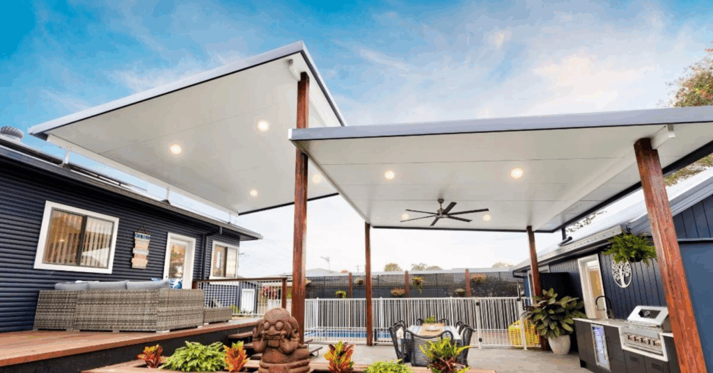 A modern garden patio with two large white-roofed pergolas featuring quality roofing for Perth patios, outdoor seating, a barbecue, ceiling fan, decorative plants, and a small Buddha statue. A fenced swimming pool is visible in the background under a blue sky.