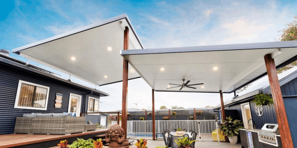 A modern garden patio with two large white-roofed pergolas featuring quality roofing for Perth patios, outdoor seating, a barbecue, ceiling fan, decorative plants, and a small Buddha statue. A fenced swimming pool is visible in the background under a blue sky.