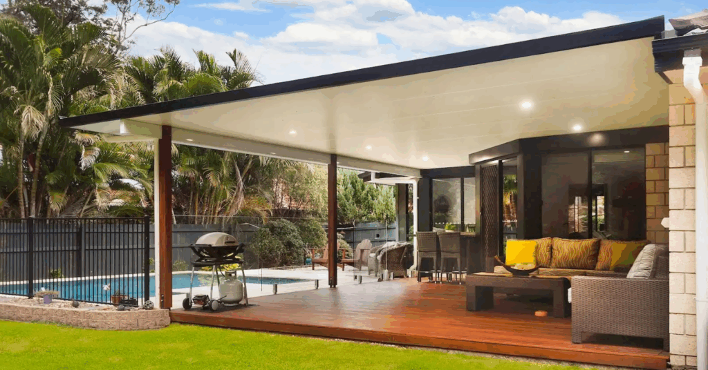A modern back garden patio with a covered wooden decking, outdoor furniture, barbecue, lush green lawn, and a swimming pool, surrounded by a black fence and tropical plants under a partly cloudy sky.