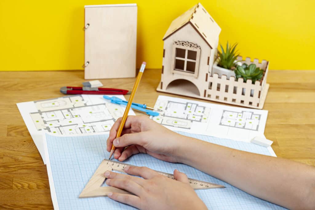 A person uses a pencil and ruler to draw on graph paper, with house plans, model house, notebook, and drawing tools on a wooden table against a bright yellow background.