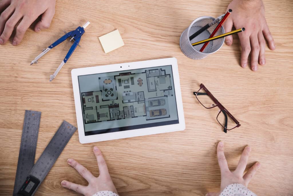A tablet displaying a house floor plan sits on a wooden table surrounded by rulers, a compass, pencils, a sticky note, spectacles, and the hands of three people engaged in discussion.