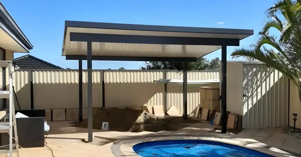 A modern metal patio cover stands beside a small round pool in a fenced back garden, with construction materials and sand piled under the shelter.