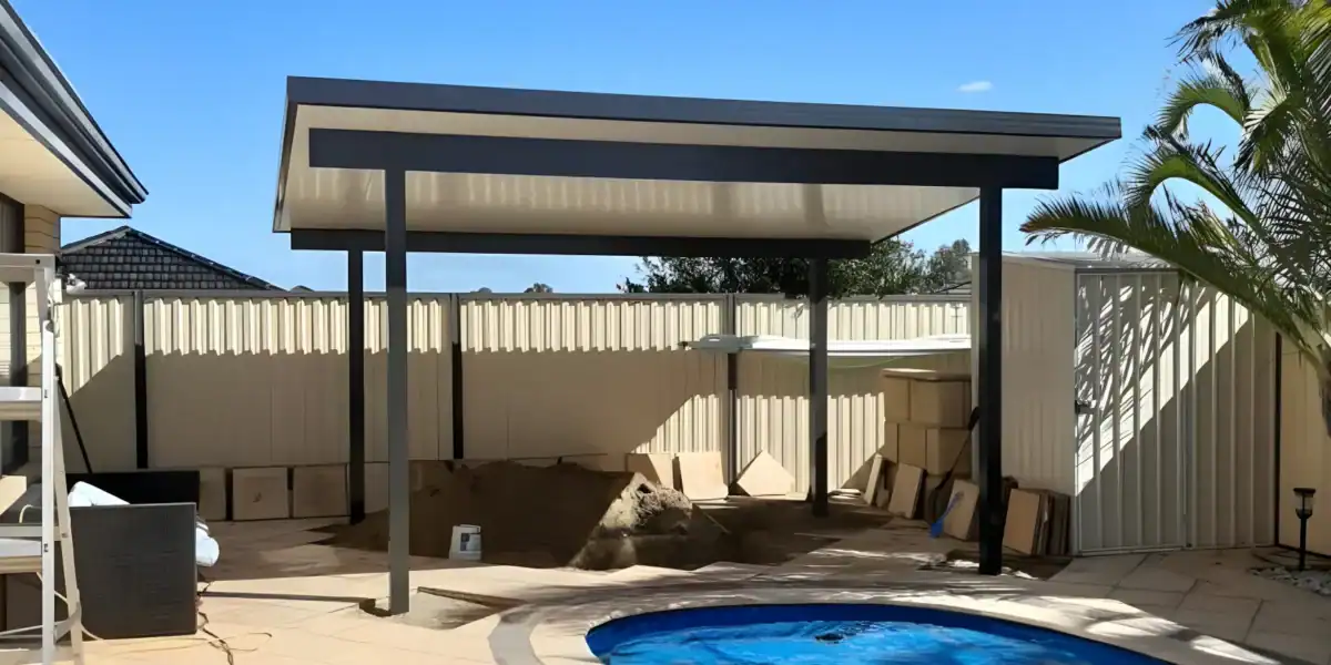 A modern metal patio cover stands beside a small round pool in a fenced back garden, with construction materials and sand piled under the shelter.