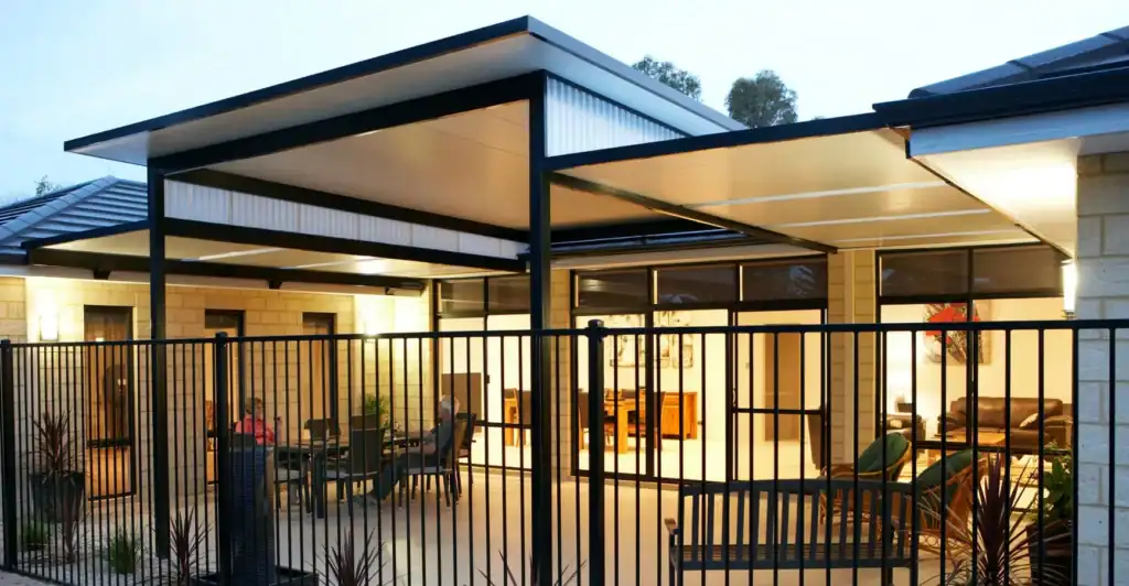 Modern outdoor patio area with a metal fence, covered seating, glass doors, and interior lights visible from outside at twilight.