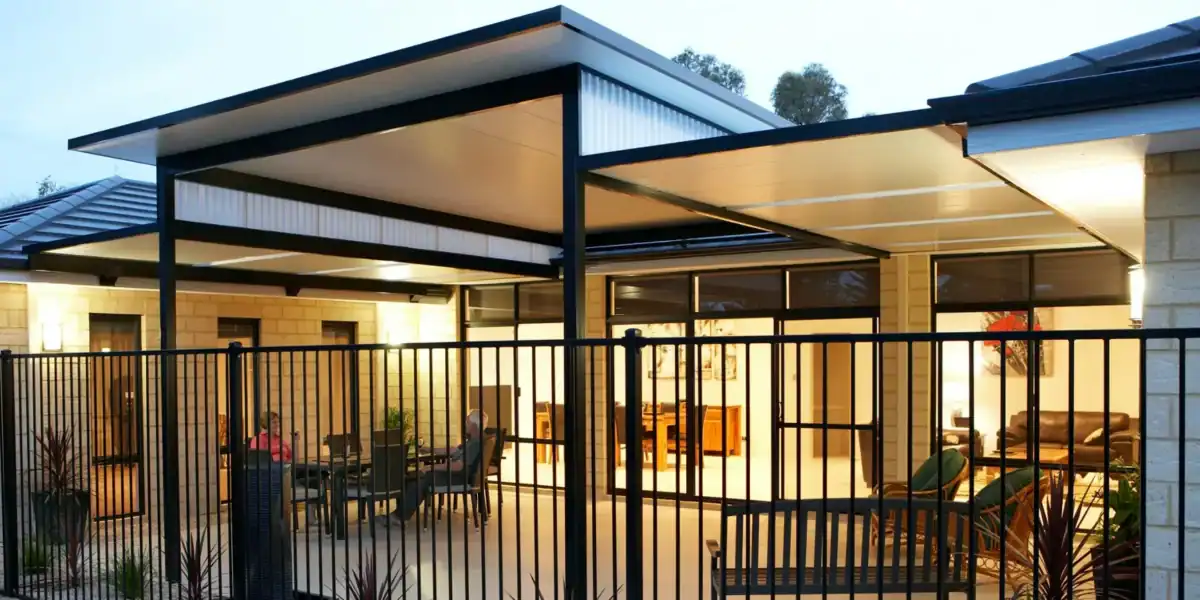 Modern outdoor patio area with a metal fence, covered seating, glass doors, and interior lights visible from outside at twilight.